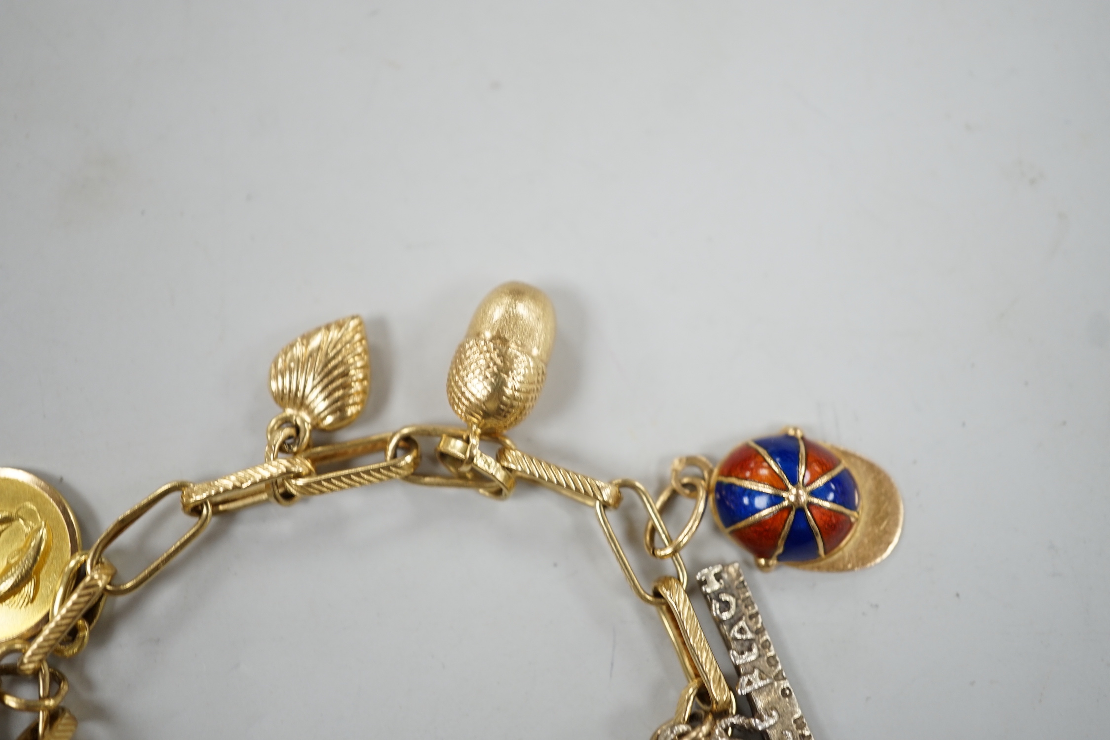 A modern Italian yellow metal charm bracelet, hung with eleven assorted charms, including 750 and enamel jockey cap, agate clasped hand and 750 acorn